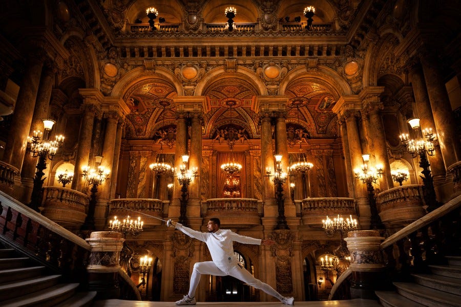 A fencer with an épée poses inside a grand opera house.