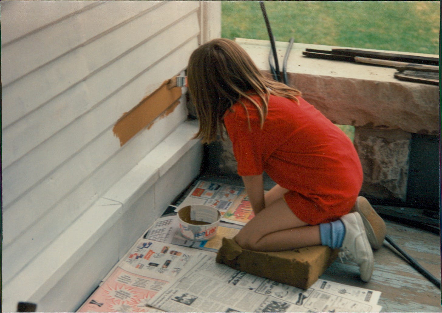 Child holding paint brush kneeling while painting the side of a house. 