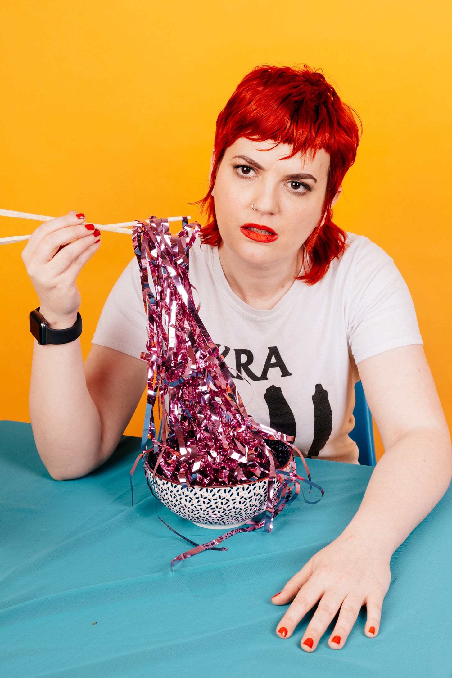 A woman with a red mullet hair style uses chop sticks to eat glittery pretend noodles