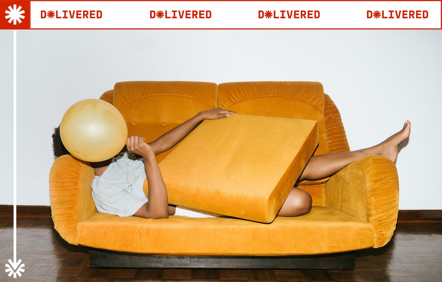 A person of colour is relaxing on a bright orange couch and covers their face with a golden balloon.