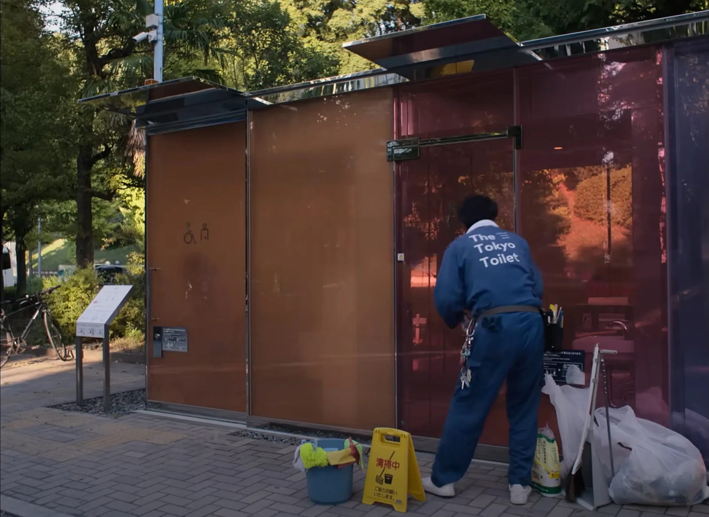 HIrayama in his blue uniform leans down to unlock the door to one of his restrooms, made up of large panes of colored glass.