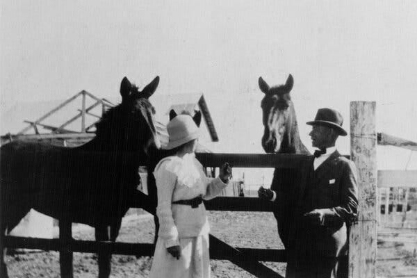 A man in a hat and suit stands with a woman in a white sweater, skirt and a hat in front of two horses in a paddock area. 