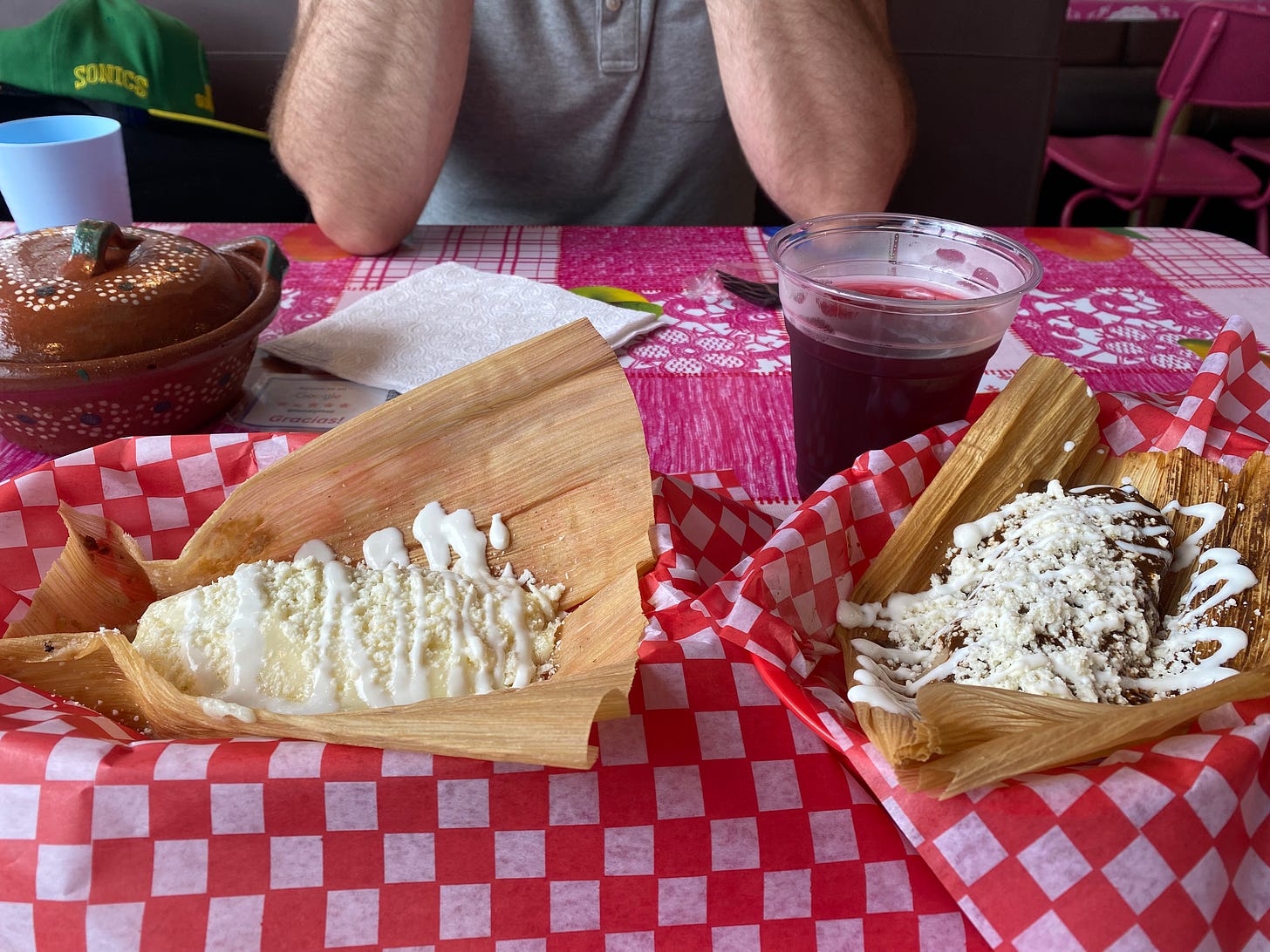 Two baskets with red and white checkered paper, each with a different tamale inside, the corn husks open and the tamales covered with crumbles of queso and drizzles of crema. Behind them is a painted stoneware pot with a lid, a clear cup of light purple agua de jamaica, and on the opposite side of the table, Iain, wearing a grey shirt, rests his elbows on the table.