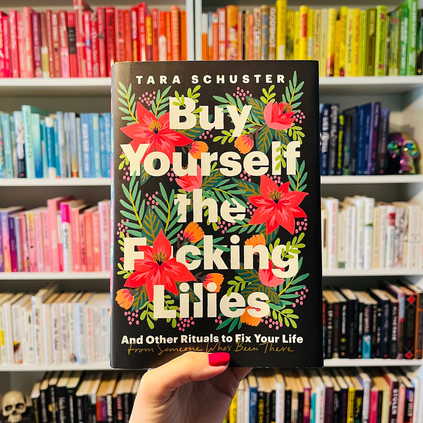 A hand holding a book, Buy Yourself the Fucking Lilies by Tara Schuster, in front of a bookshelf.