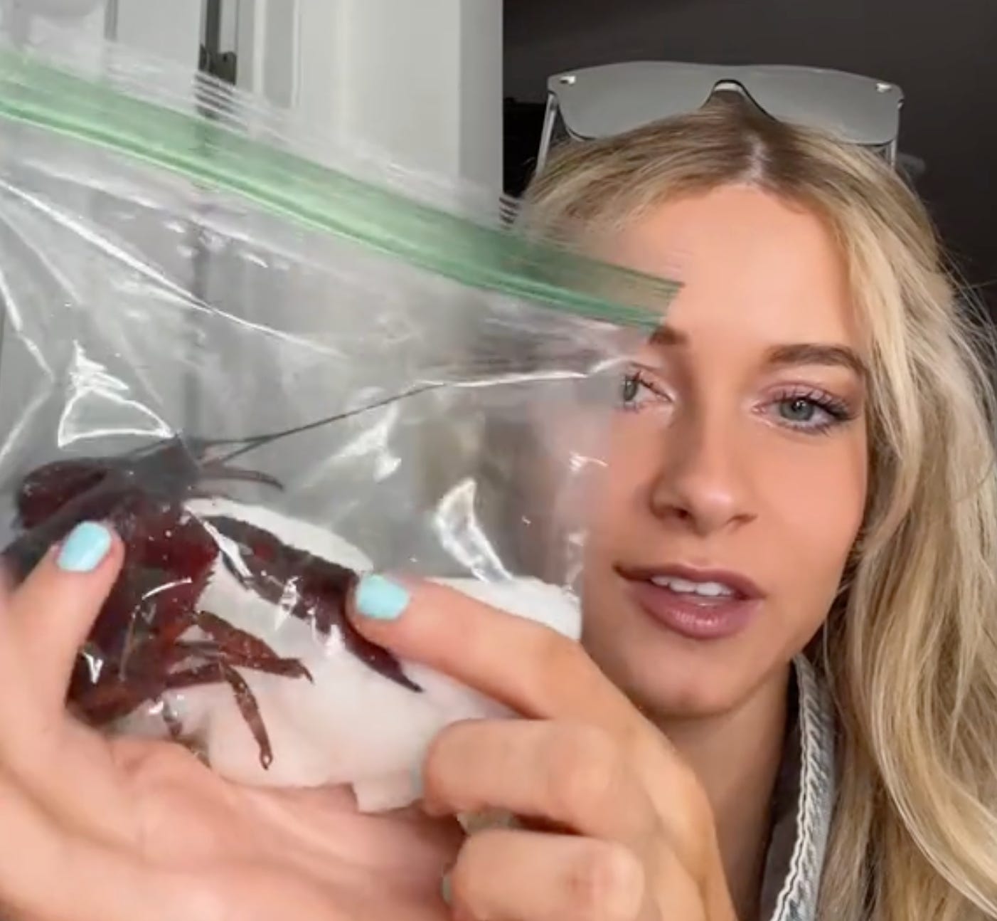 A young blonde woman holds up a ziploc bag containing a wet paper towel and a live crawfish.