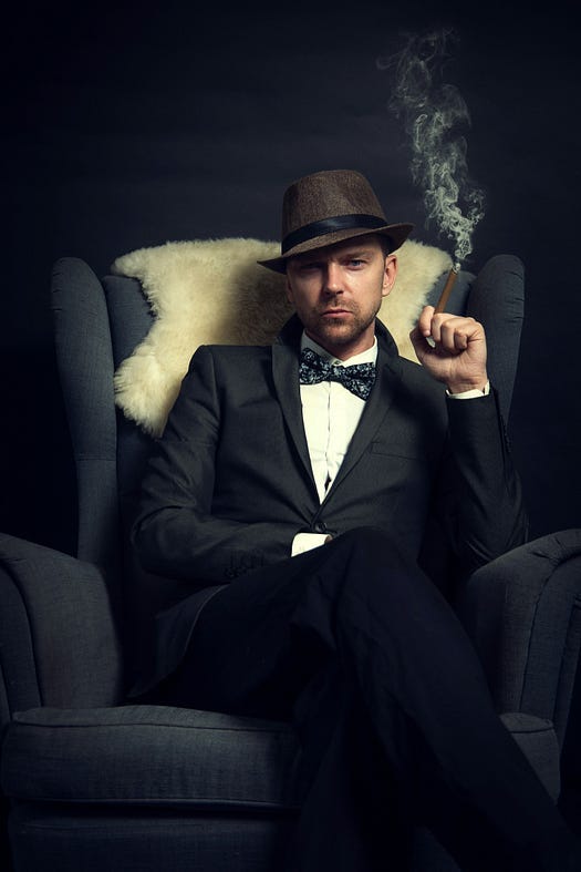 Man with hat and suit sitting on a chair with a cigarette in hand.