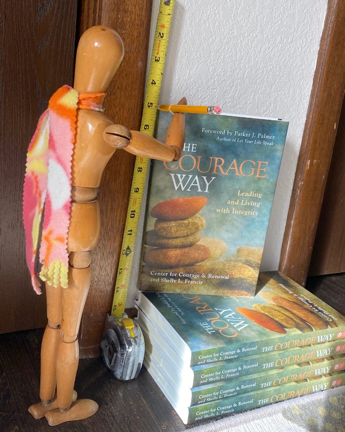 An art manikin or Emotikin measuring the height of five copies of The Courage Way, marking the height on the wall with a pencil. 