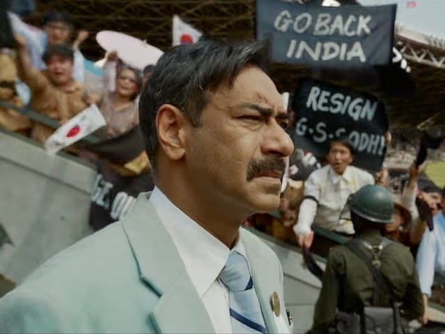 A still from Hindi film, ‘Maidaan’. Bust of S A Rahim, played by Ajay Devgn, in a greyish-blue suit and a blue neck tie as he looks intently into the distance. The background has protestors holding banners. 