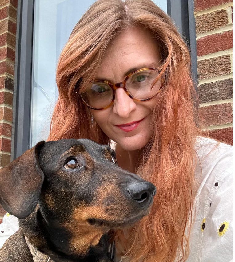  Rachel sits before a brick wall. She is white and has long brown/red hair. She wears glasses. She is holding her brown sausage dog, Rusty. 