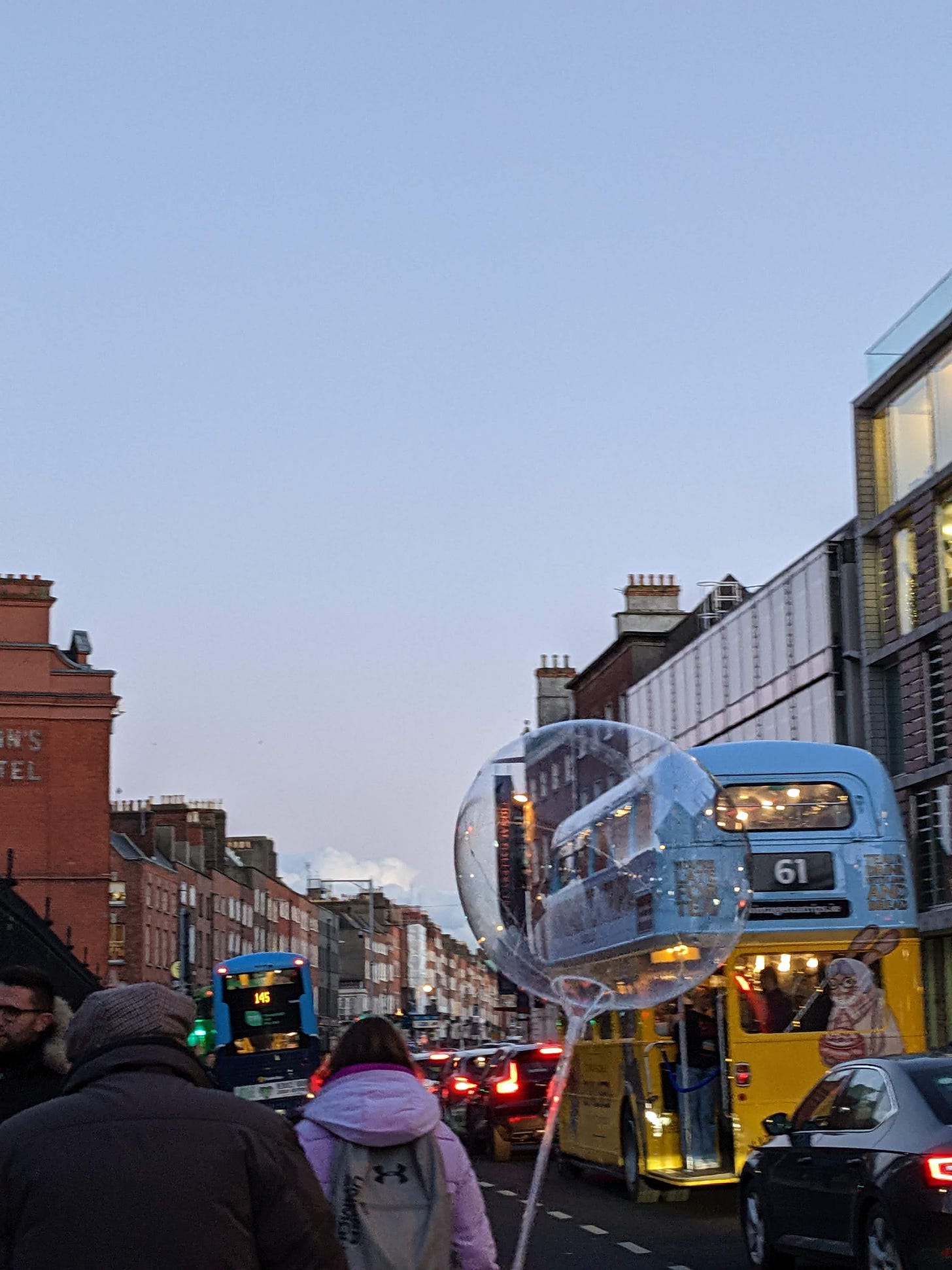 a busy city street—buses, cars, buildings—at winter dusk. in the foreground a child walking away from the camera carries a transparent, iridescent balloon that is at the center of the image and distorts the view through it very slightly.