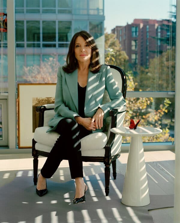Marianne Williamson, wearing a light teal blazer over a black shirt and black slacks, sits on a cream-colored chair in front of a window in her home.