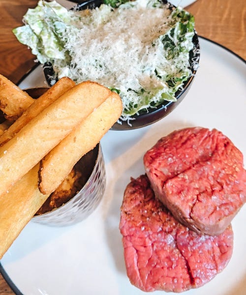 A white plate with steak, a silver bowl of chips, and a caesar salad on it