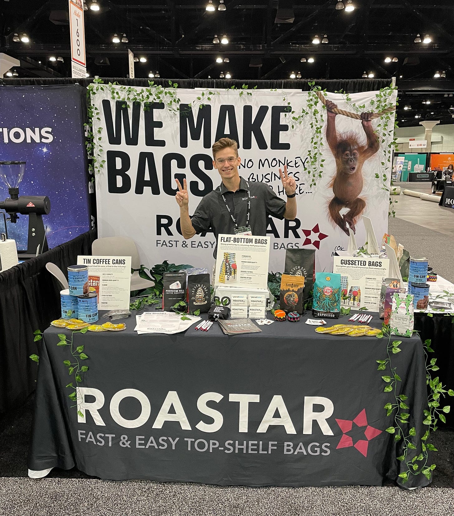 A salesman flashes the peace sign with his fingers while standing behind a table loaded with sample coffee bags and merchandise at a trade show.