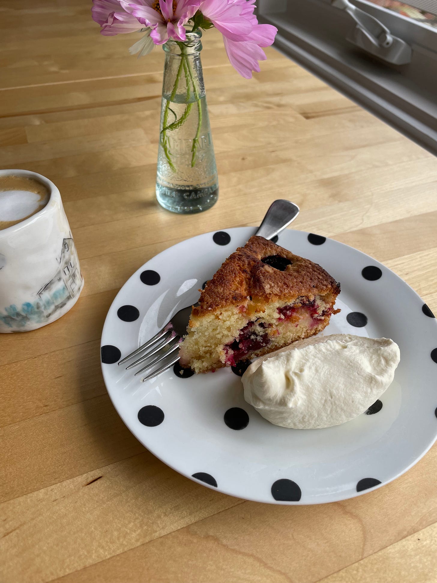 Slice of butter cake with sunken plums on a spotty plate, with whipped cream served alongside. A small flat white and vase of flowers are also on the blonde wood dining table.
