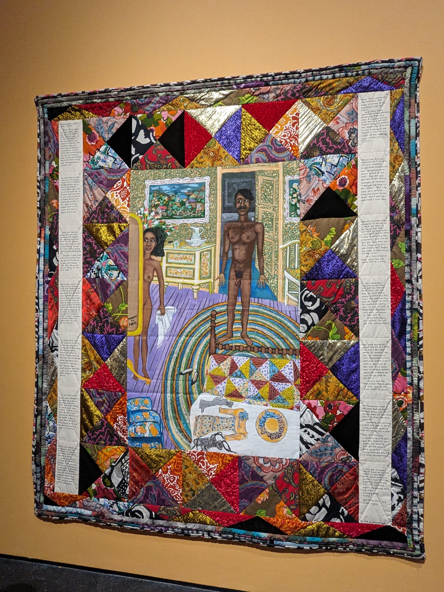 A quilt haging against a yellow wall. Diamond shaped prints run along the quilt next to text written by the artist. In the center is a naked Black man standing in front of a Black woman 