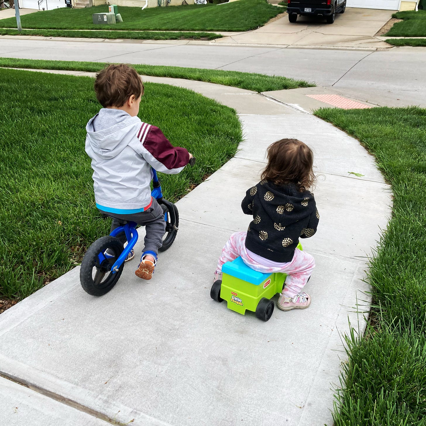 The picture shows two children on a sidewalk between areas of green grass. The three year old boy is on a balance bike and the two year old girl is on a ride-on toy train. They are facing away from the camera.