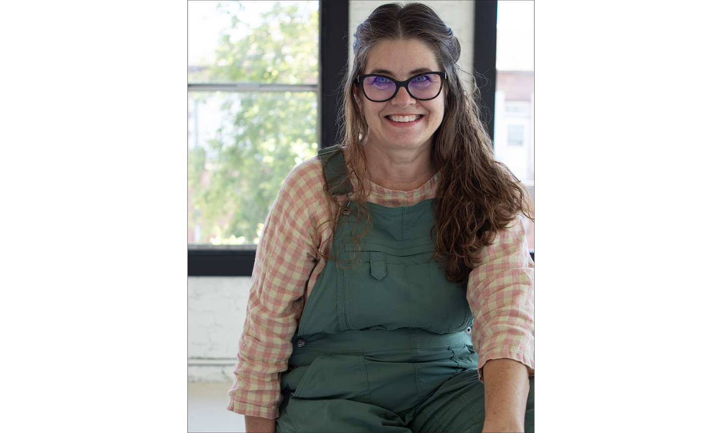 Anne, a white woman, is smiling at the camera. She's wearing green overalls and a pink gingham handmade shirt.