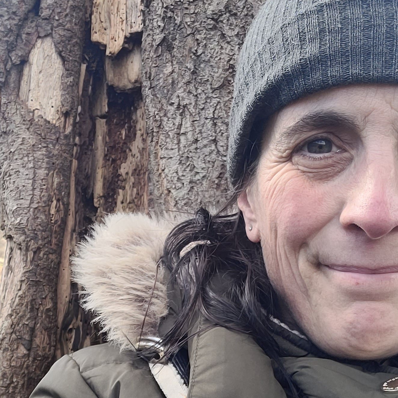 A woman in a winter coat and grey hat sits in a tree. She has taken a selfie but it is only showing half her face. She is smiling, but her eyes look sad