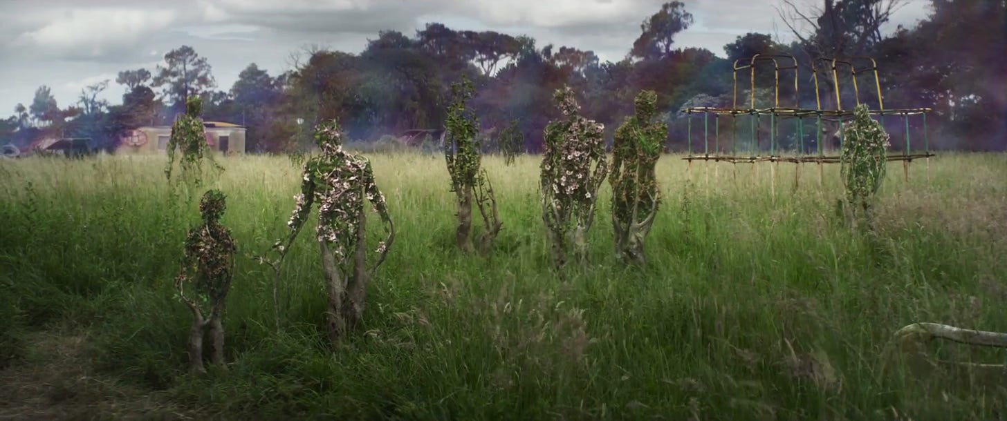 Image is a field of flowering woodsy shrubs in the shape of humans standing in a field of tall green grass. In the distance is a stripped wooden structure, dilapidated houses, and a forest of deciduous trees.