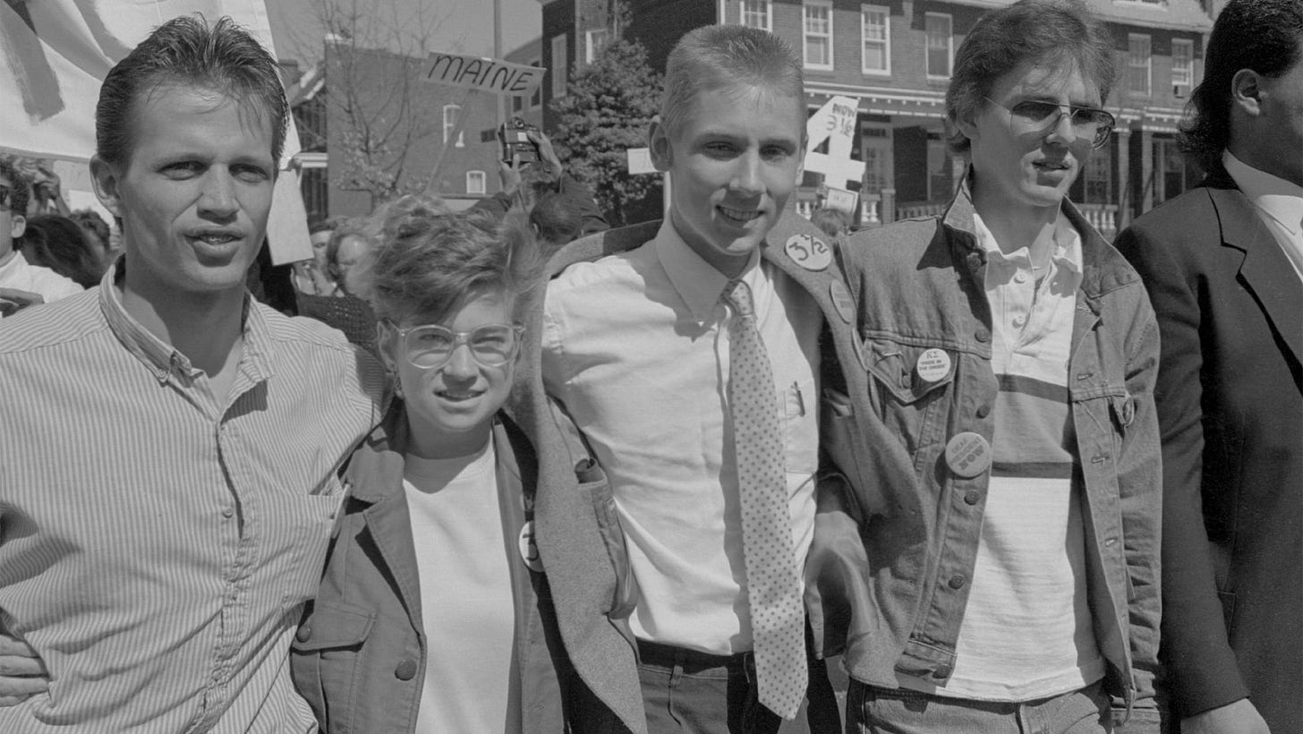4 white Deaf students wear some classic 1980s clothes as they embrace for a black and white photo.