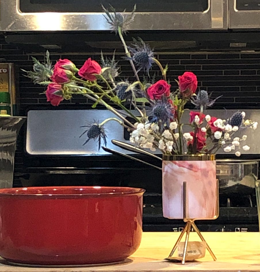 A bouquet of red roses, blue thistles, and white baby's breath in a small pink vase. You can also see a red bowl and an oven in the background.