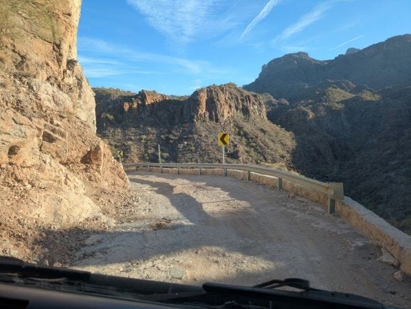 a heavily rutted dirt road on the edge of a cliff, winding around a tight bend with a flimsy guardrail and a yellow arrow warning sign