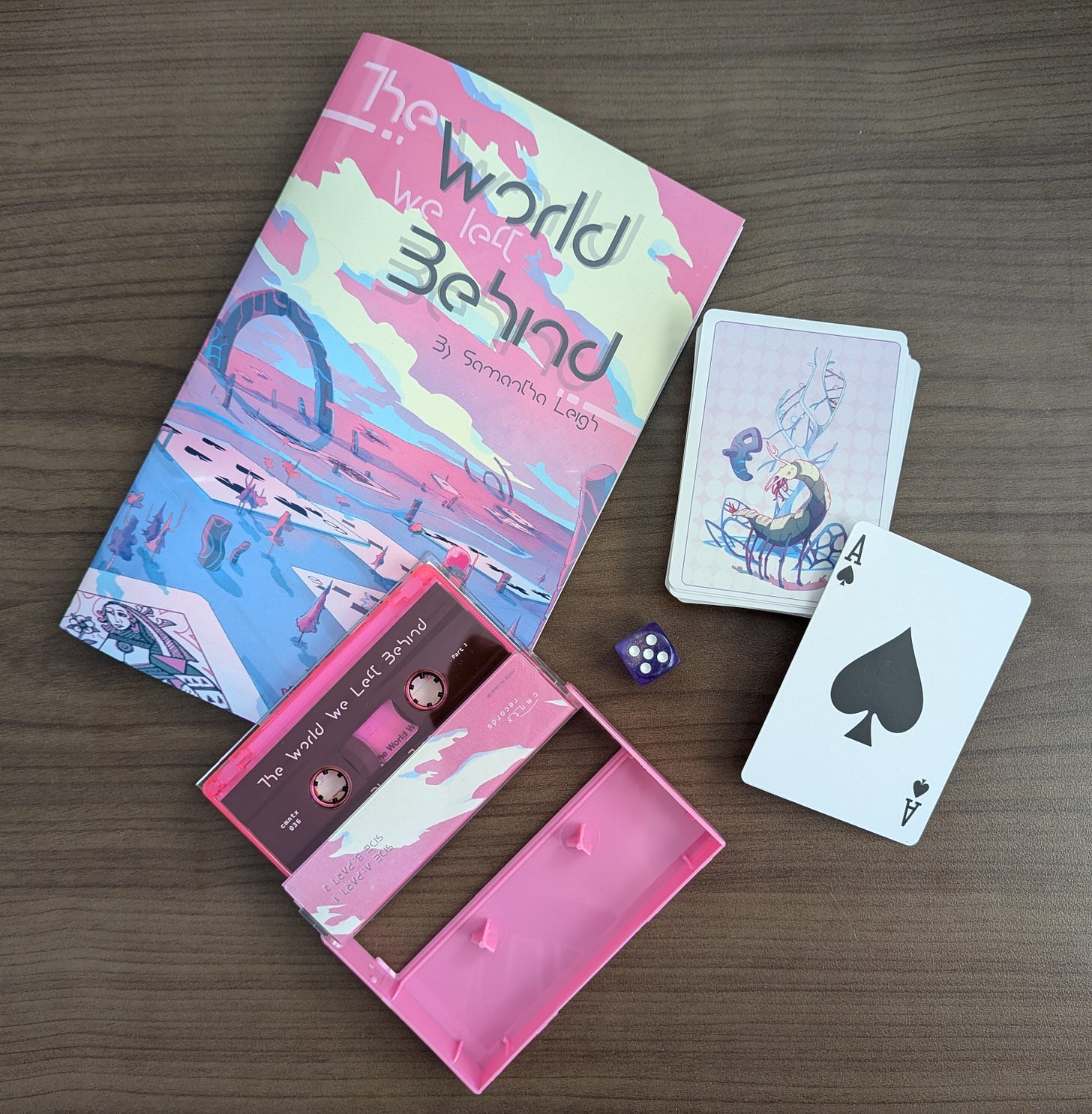 A copy of The World We Left Behind TTRPG, a cassette, a purple shimmery die, and a deck of playing cards with art from the TTRPG on the back of the cards.