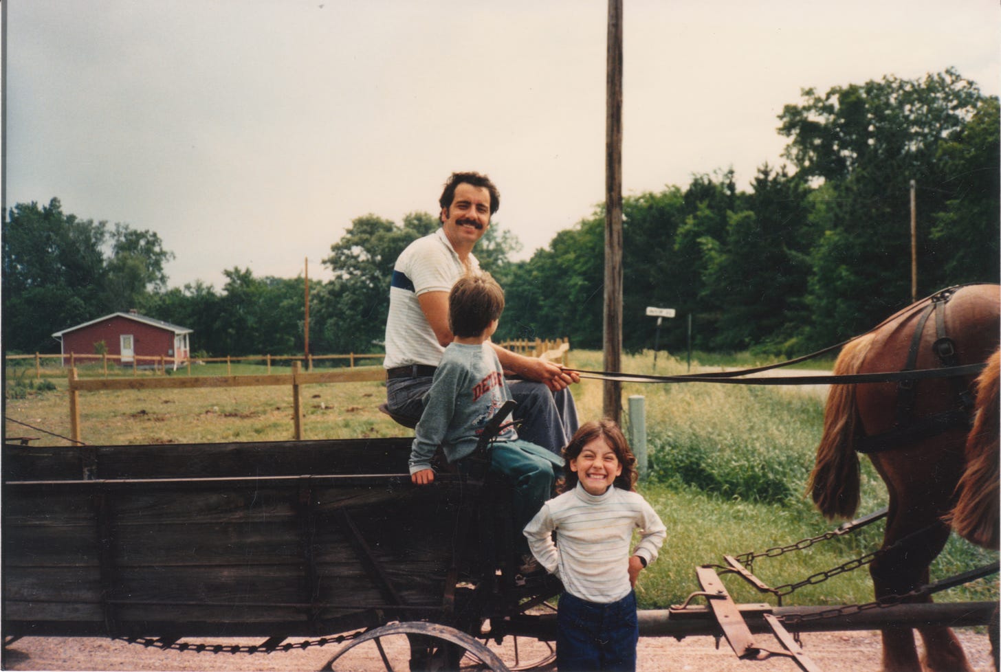 a dark-haired man with a mustache in a short sleeve white shirt sitting on an old wooden manure spreader, holding the reins of a pair of auburn Belgian workhorses. a young child sits next to him turned away from the camera. another child faces the photographer with a big smile.