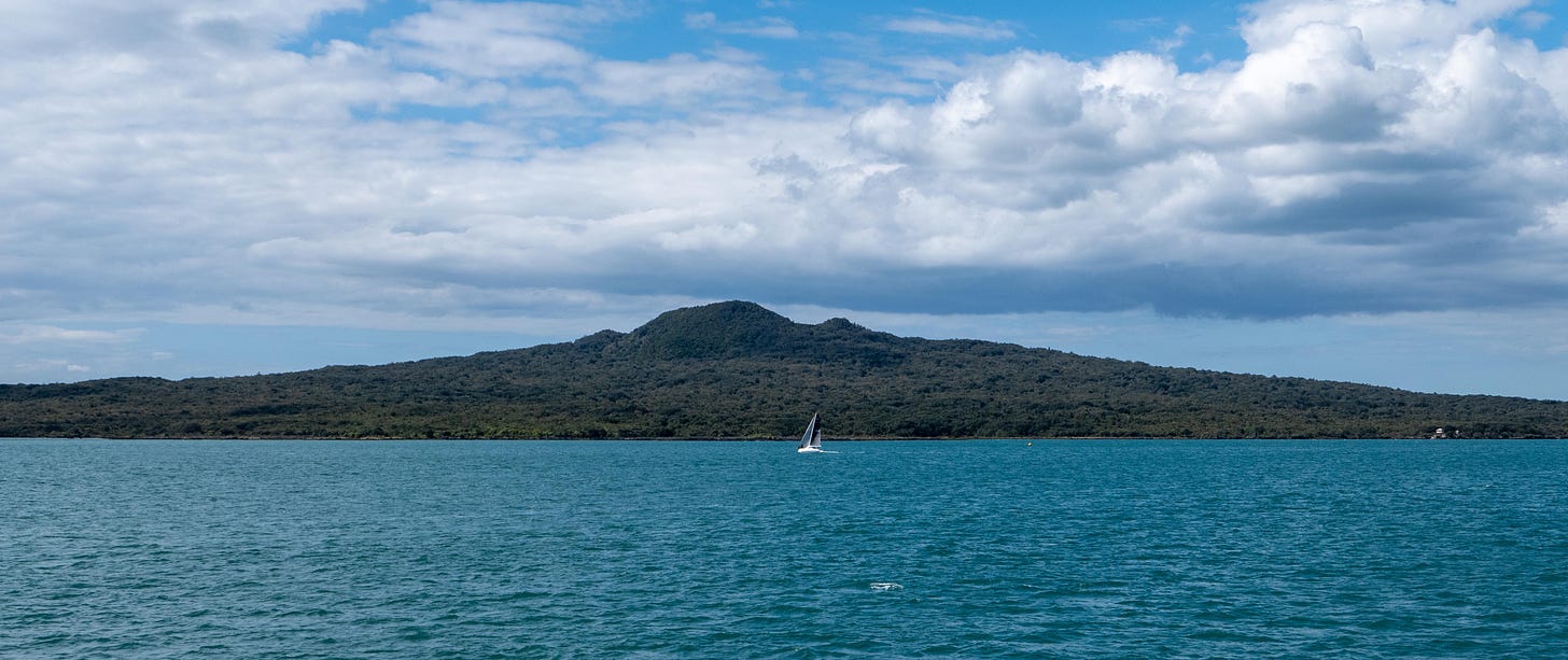 Rangitoto rising from a blue sea with a sail boat passing in front.