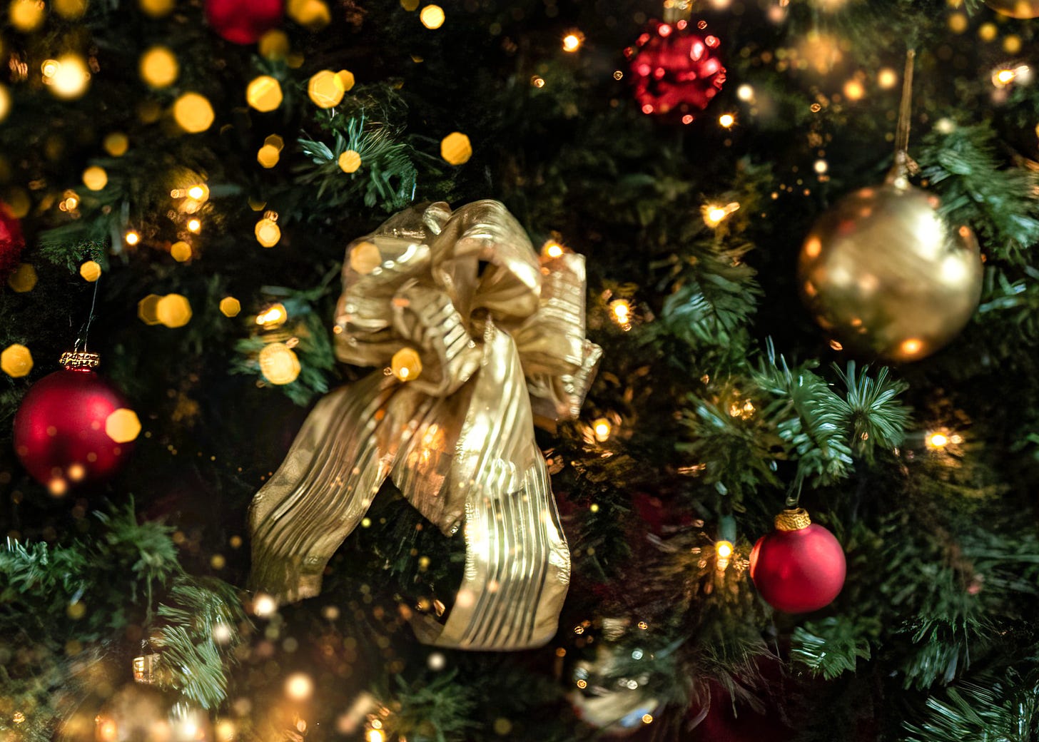 A gold bow on a green Christmas tree with twinkling lights and red and green ball ornaments