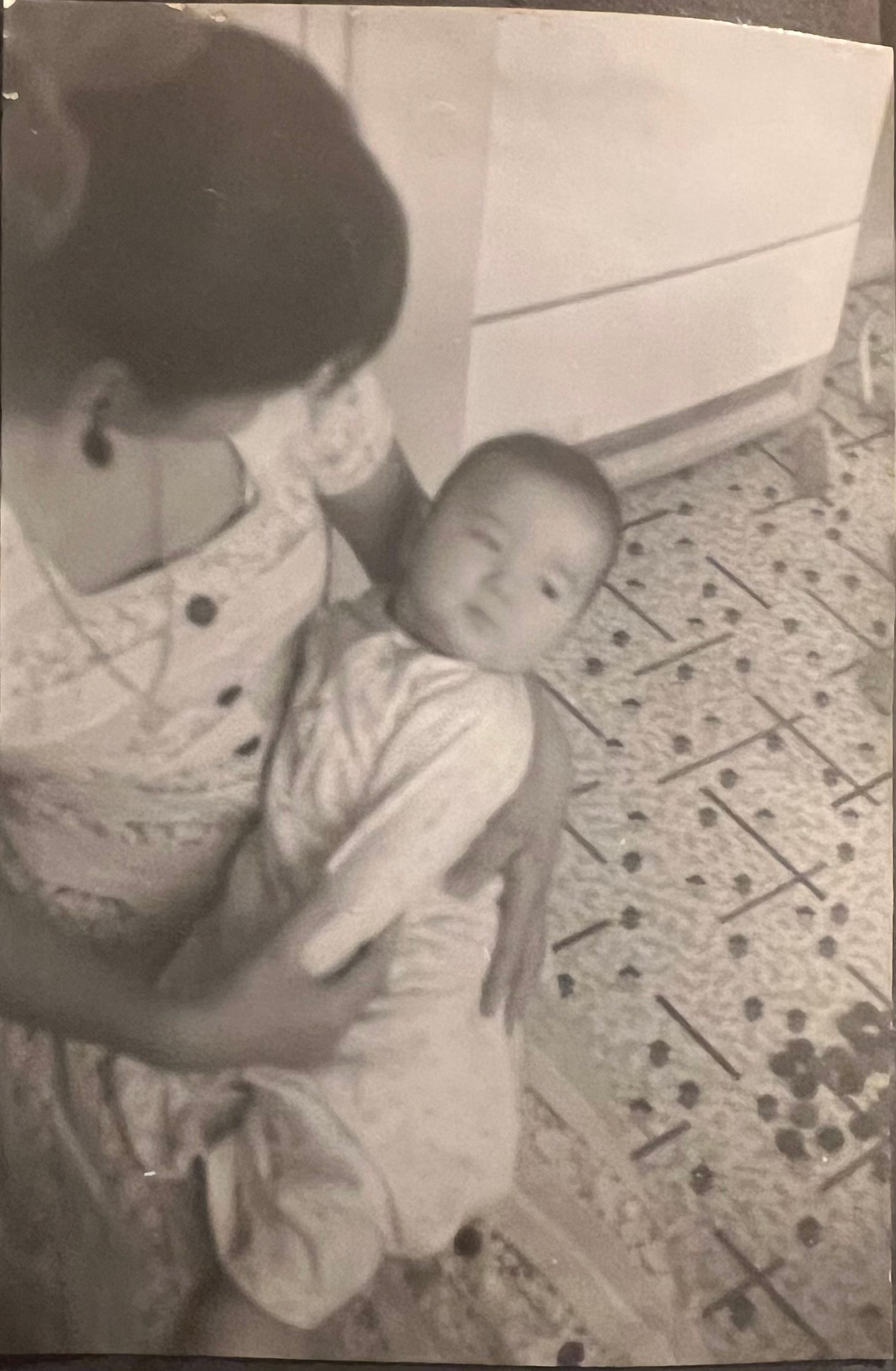 My mother holding infant me in her arms. She's looking down at me, I'm looking away. 