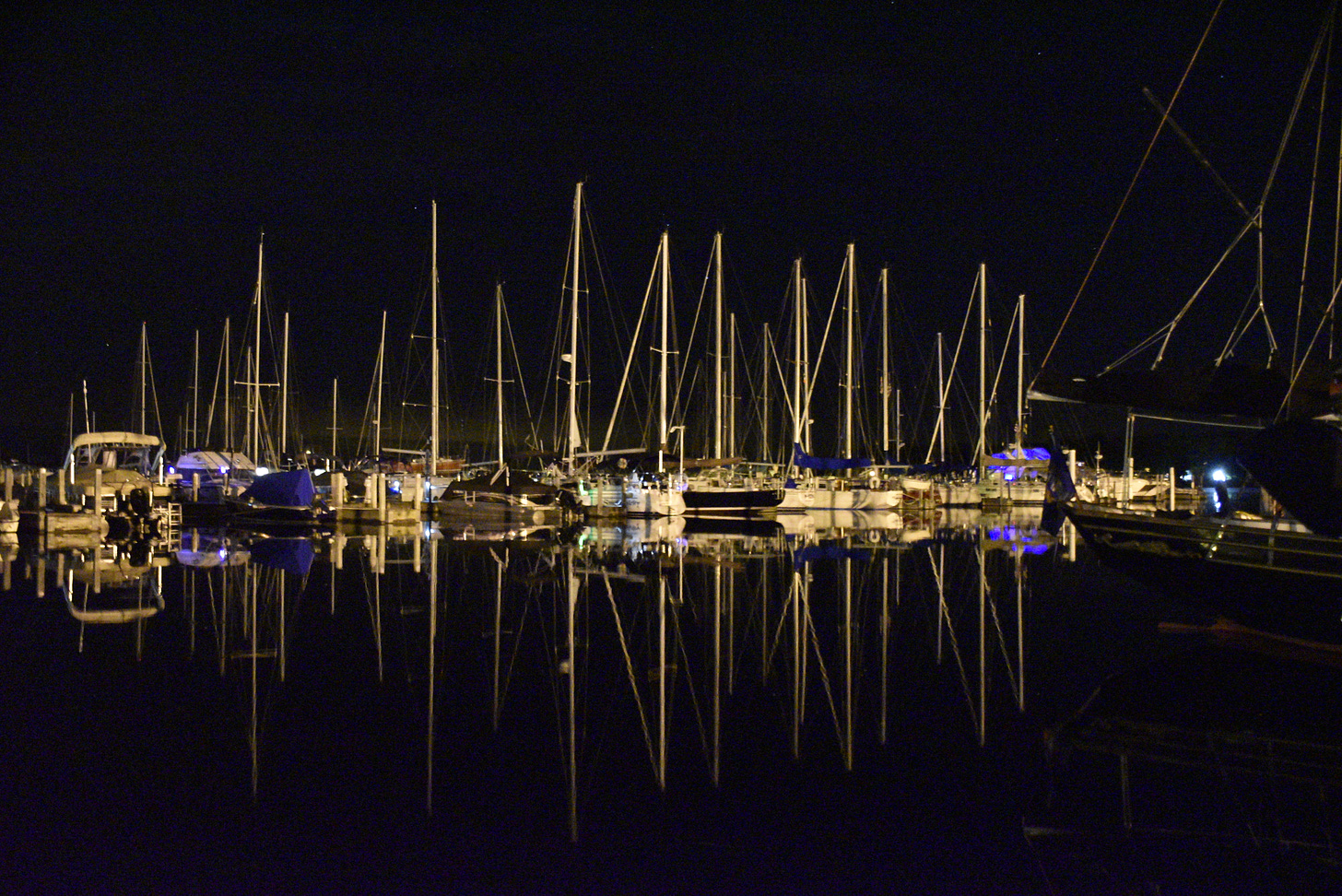 Photo of sailboats a dock at night with light reflecting images off of the water.