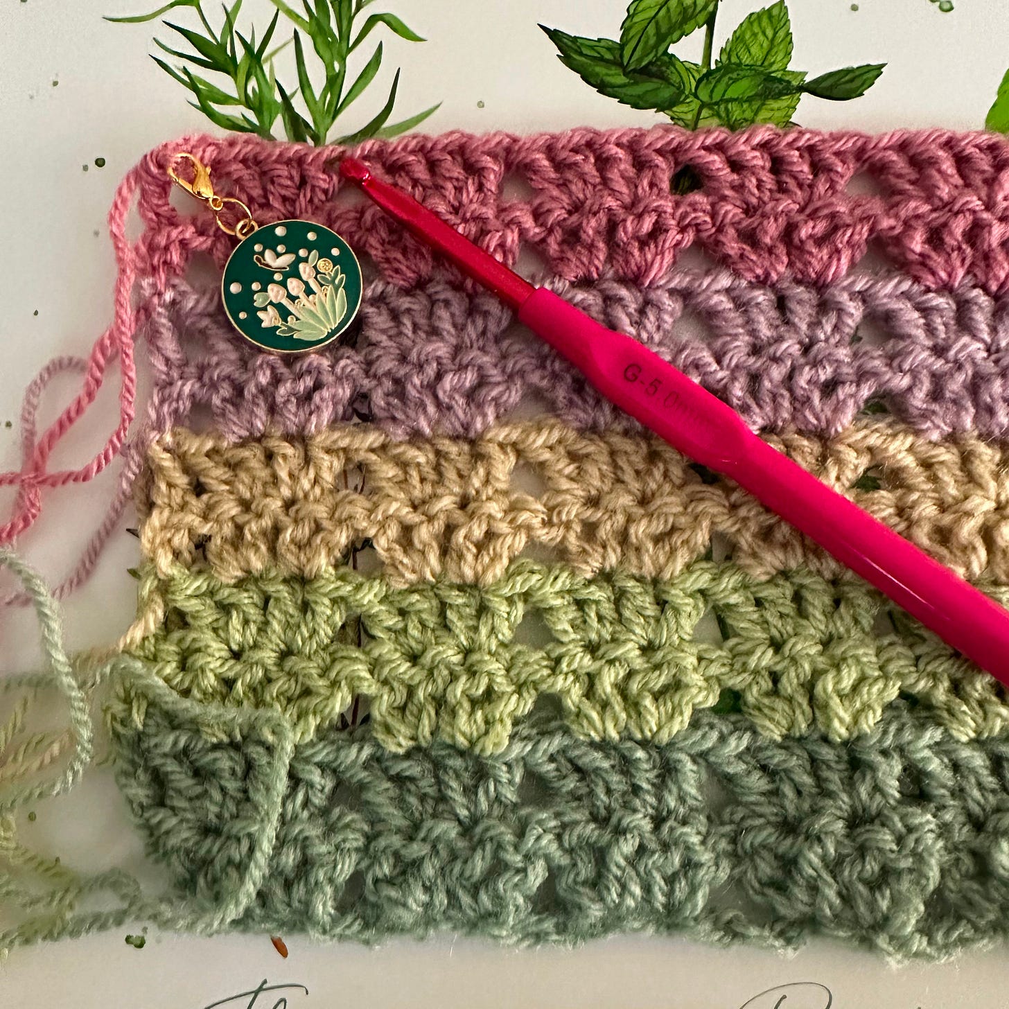 Photo of five stripes of different coloured pastel yarn, crocheted into clusters. A pink crochet hook sits on top along with a mushroom stitch marker.