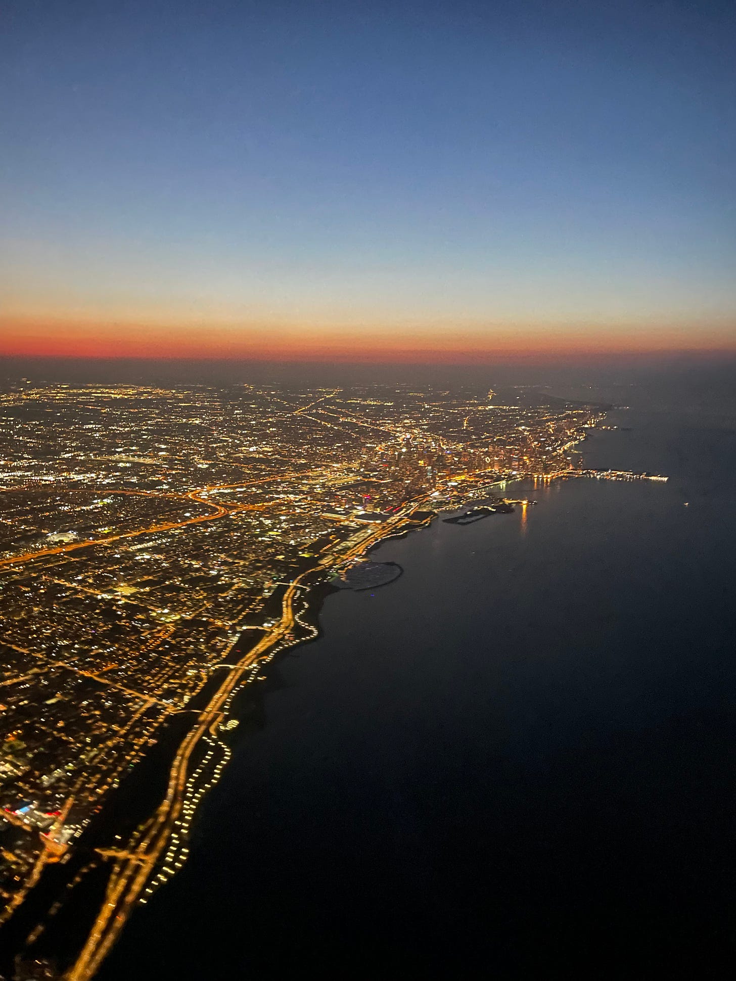 The waterfront of Chicago, seen from the air at twilight. Twinkling city lights can be seen below, and above the sky is painted hues of orange red and darkening blue.