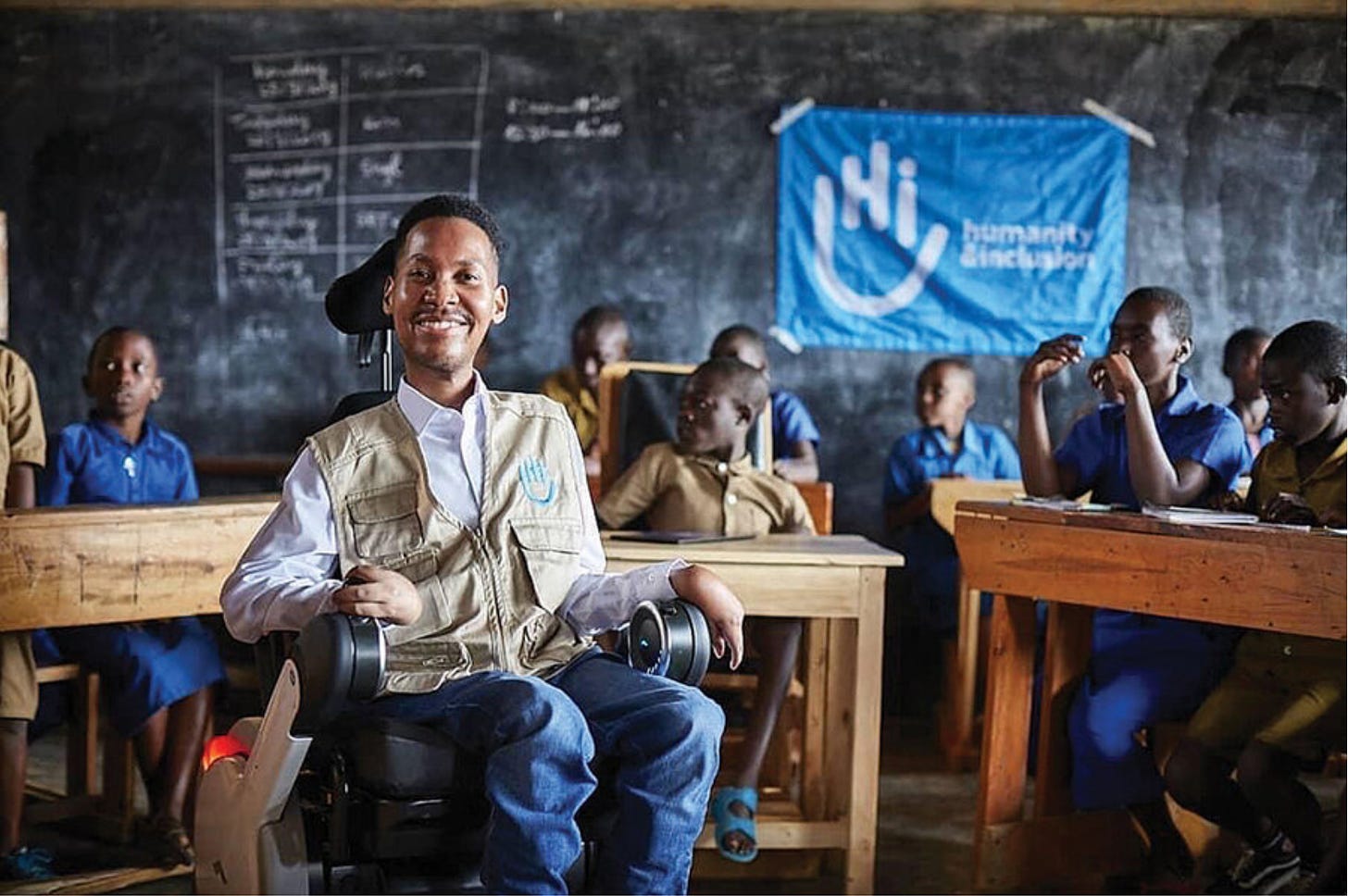 A young black man smiles from his wheelchair with a classroom of black student behind him.