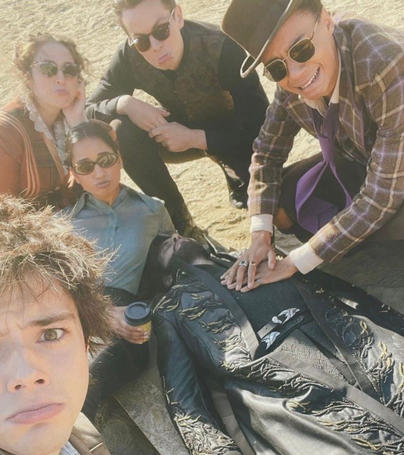 A concerned-looking Six of Crows cast in costume gathered around Ben Barnes, who is lying on the sand apparently dead. Kit Young (Jesper) has his hands on Ben's chest as though performing CPR.