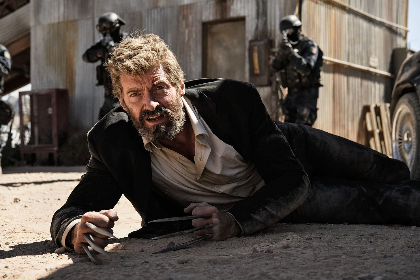 Screenshot from Logan with Wolverine lying in the dirt, claws bared, surrounded by gun-toting men in body armor.