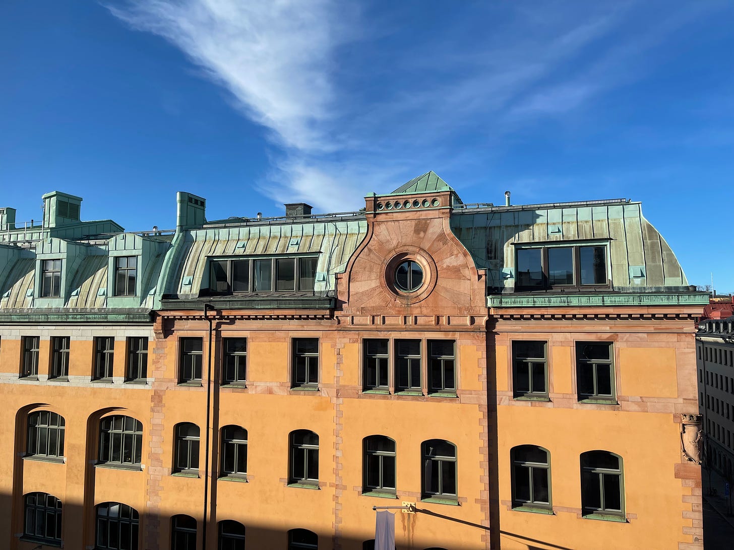 An 1800s building in yellows and pale orange with a light green metal roof, with windows in the roof and rectangular windows at regular intervals from a 6th floor window. Blue sky and a wispy white cloud above the building