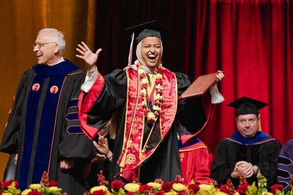 Asna Tabassum, wearing a red sash, lei, black graduation gown and black graduation cap, gives a big smile while holding a brown diploma. Professors are nearby in robes.