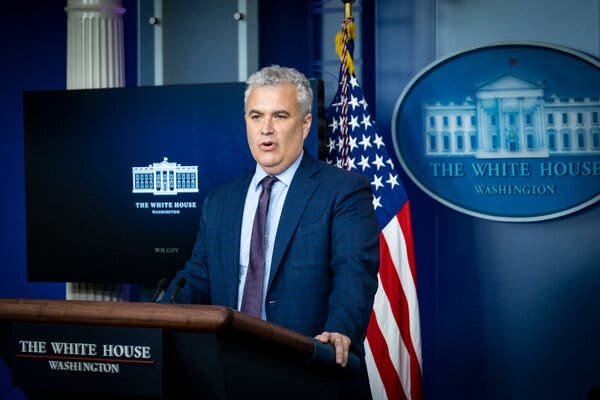 Jeff Zients speaks at a lectern with the White House emblem and an American flag displayed behind him. He is wearing a blue suit with a purple tie.