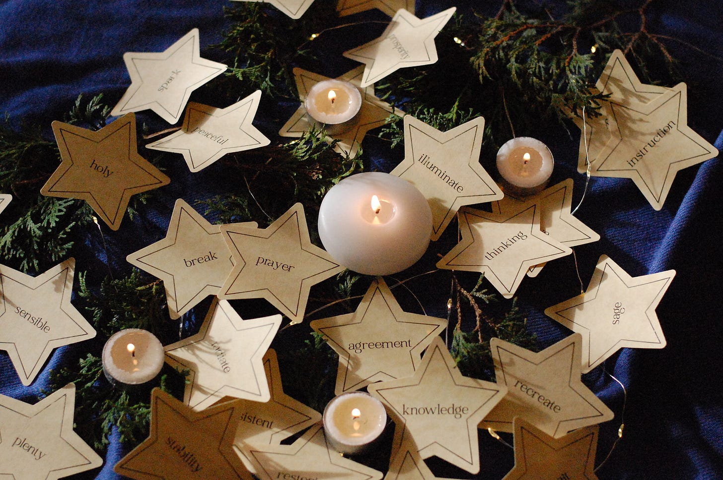 Gold paper stars, each printed with a different word, are spread out against evergreen cuttings and a blue piece of fabric, with lit tea lights scattered amongst them.