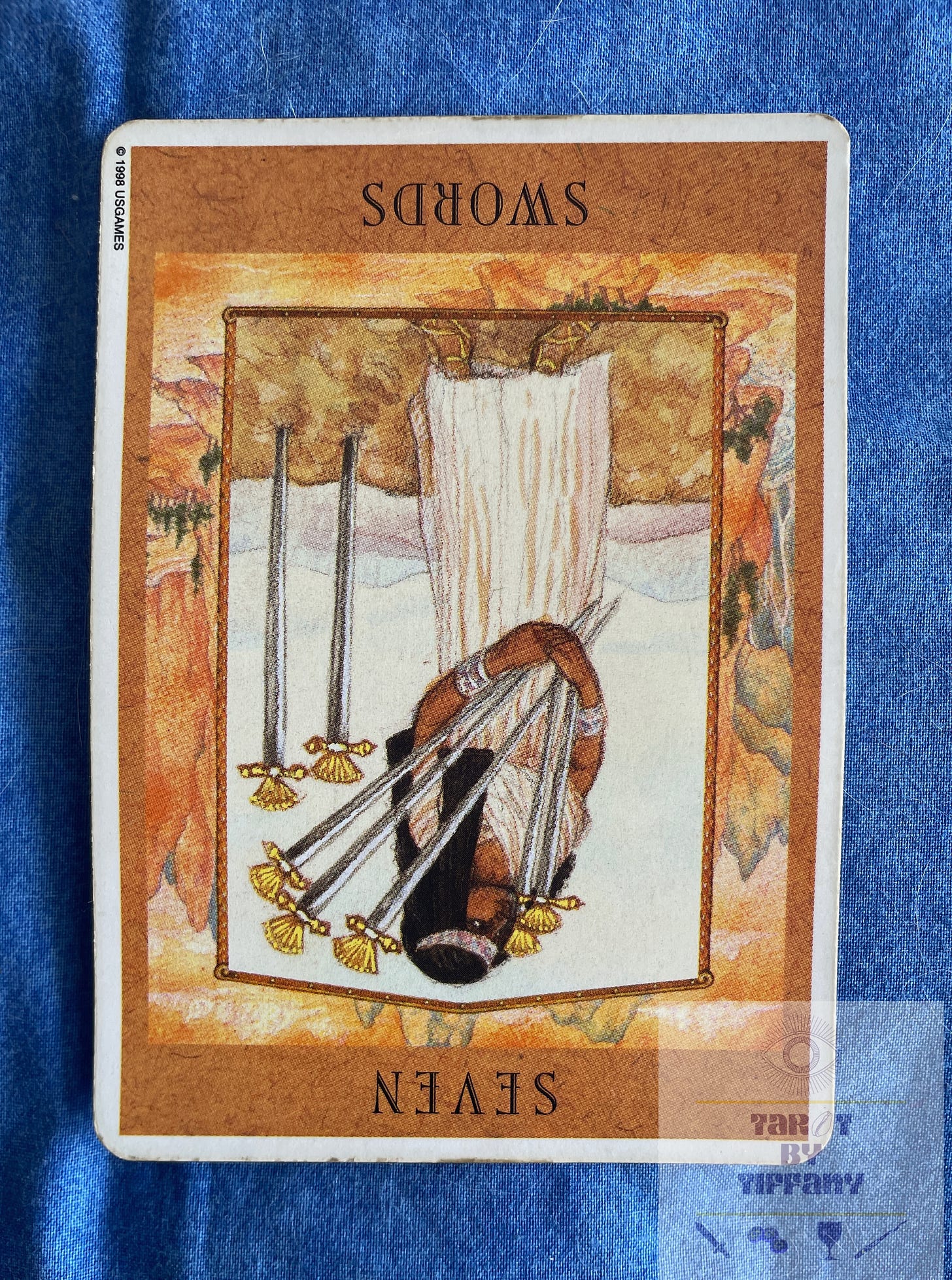 Image of the seven of swords (reversed) card from the Goddess Tarot. The card is on a blue cloth background. The card shows a brown-skinned woman in a long white gown with a white headpiece across the forehead of her long black hair. It looks Egyptian in tone. She holds five swords while two swords are stuck in the earth behind her.