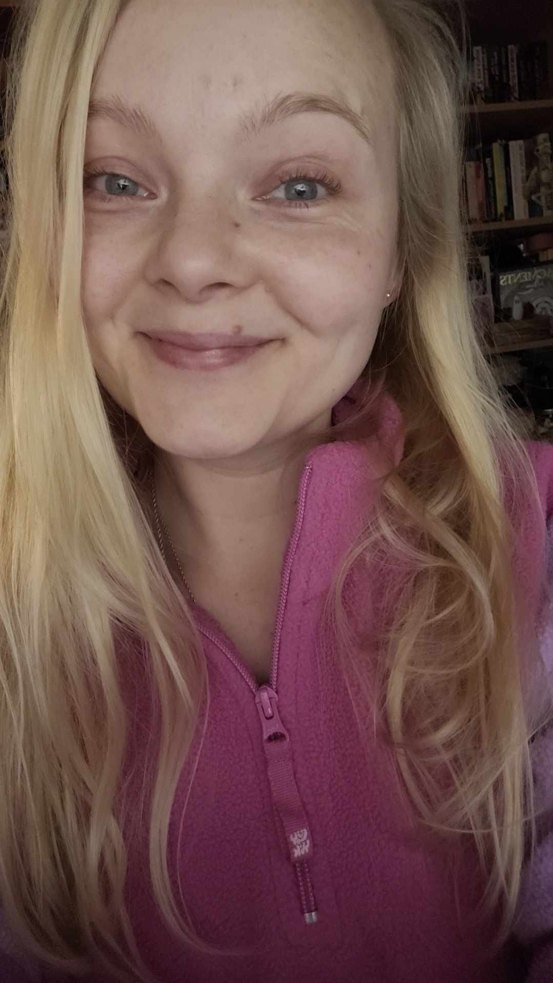 a blonde haired, white female, with blue eyes and a smiling face. She has long hair and is wearing a pink zip up fleece.