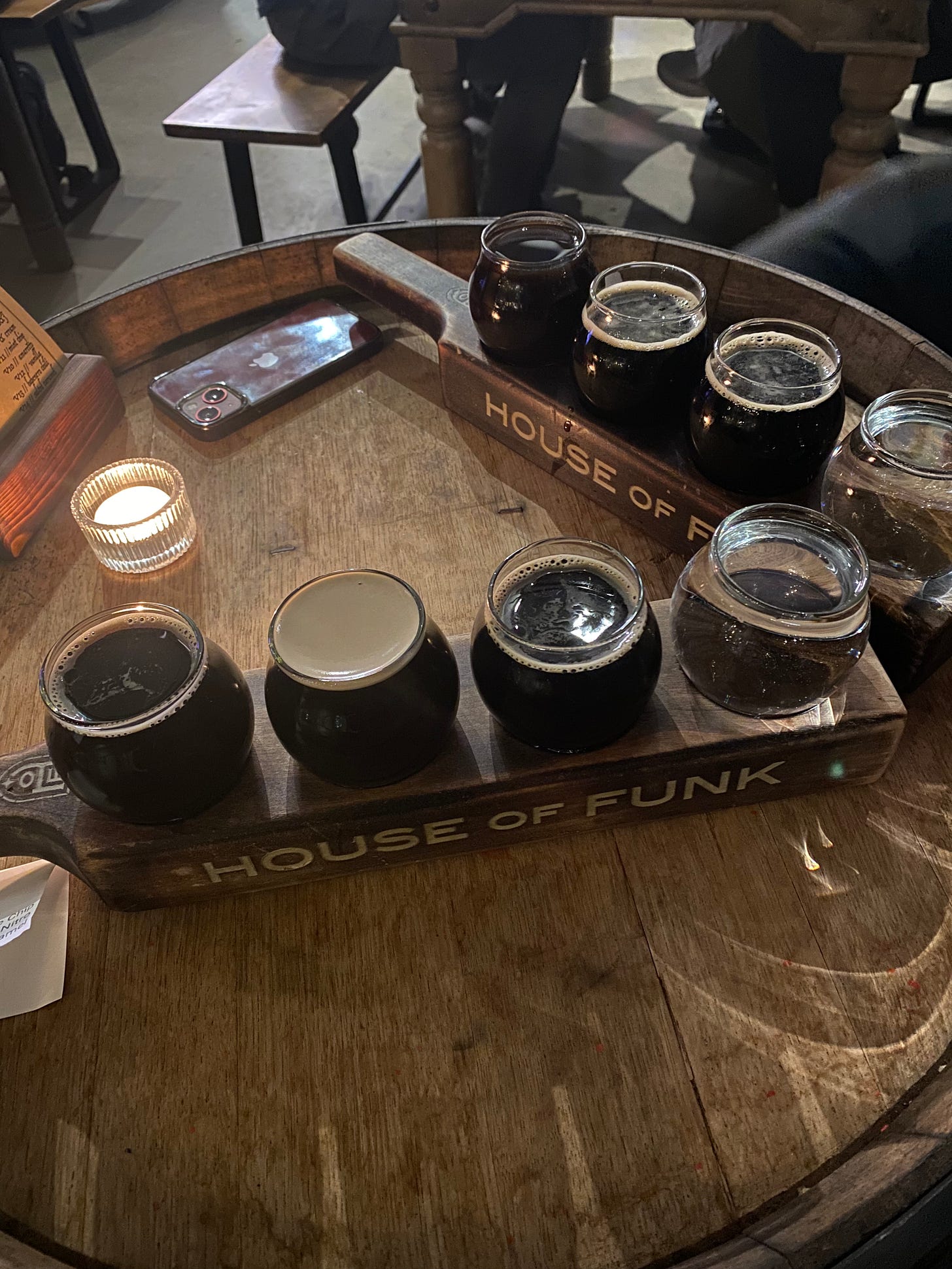 Two wooden beer flight paddles emblazoned with House of Funk lettering, each with a flight of three dark beers and a fourth glass with water. The paddles are on top of a table made from a barrel which has a tealight and a menu in a wooden stand sitting on it. Other tables and benches are visible in the background of the photo.