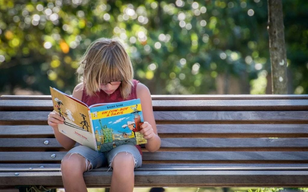 A young girl reads a book that she got from a little free library on a park bench