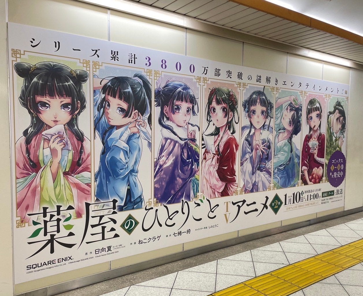 Large billboard attached to the wall of a train station corridor, lit from above with fluorescent lights. The billboard displays eight color illustrations of "The Apothecary Diaries" protagonist Maomao.