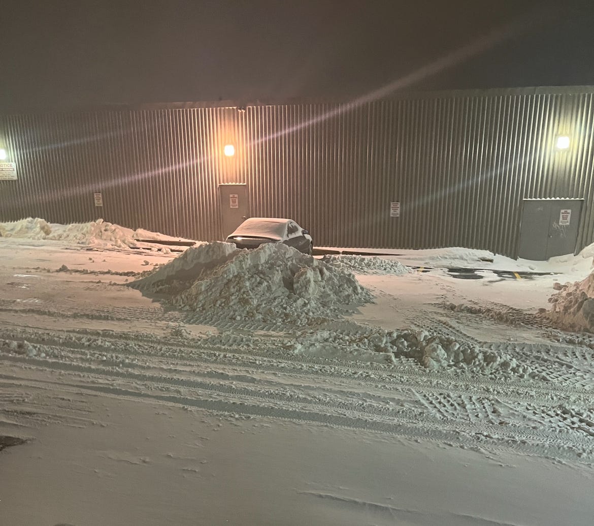 A car is parked in a snowy parking lot agsinst the side of a building. a snow plow has piled a bunch of snow right behind the car so the driver couldn't leave if he wanted to.