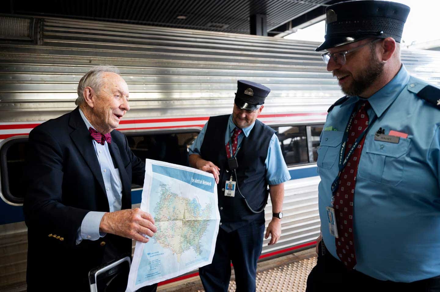 Older man holds an Amtrak map next to a train in the station while speaking with two Amtrak conductors.