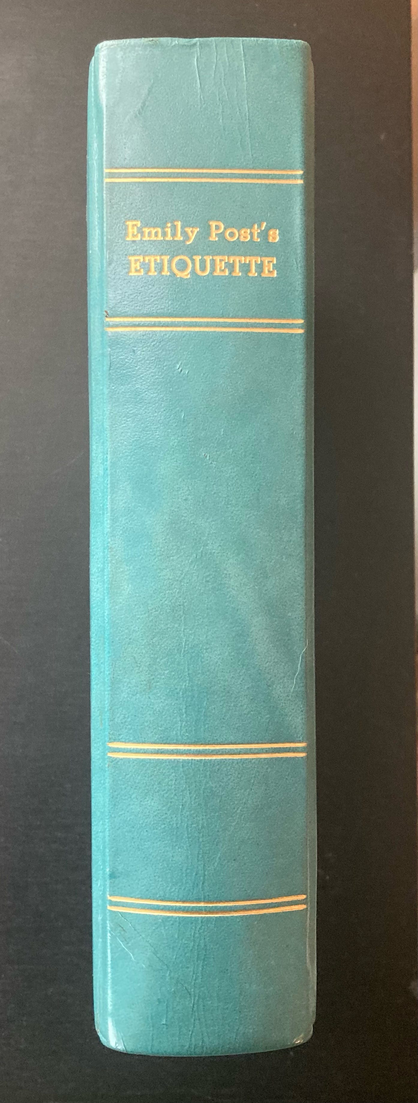 black background, teal leather book spine with gold lettering Emily Post's Etiquette and a few gold lines to help with spacing
