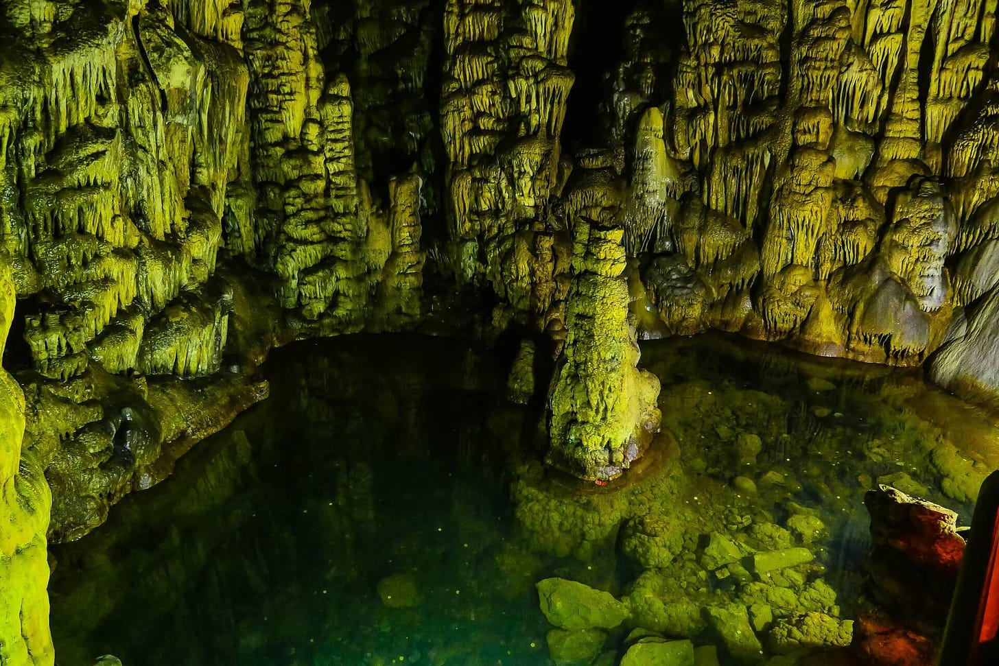 Foto do interior de uma caverna, mostrando várias estalactites e estalagmites, formações rochosas que se acumulam, como se derretessem lentamente, em tons esverdeados.